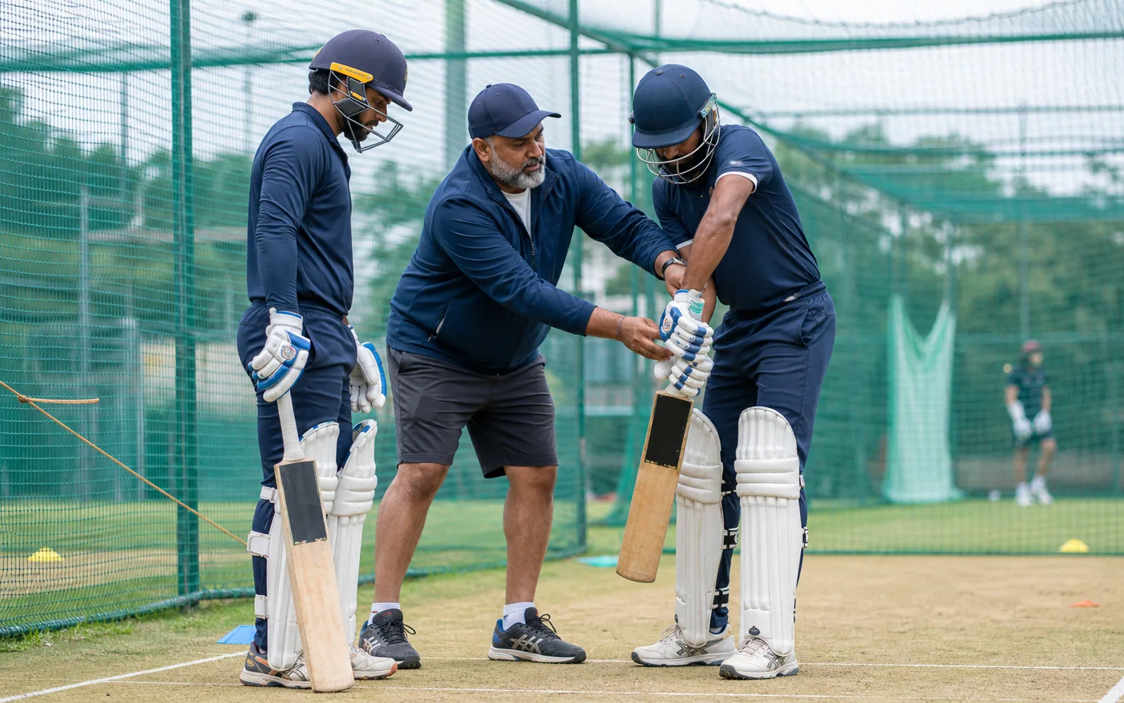 A cricket coach guiding two players through batting technique in the nets